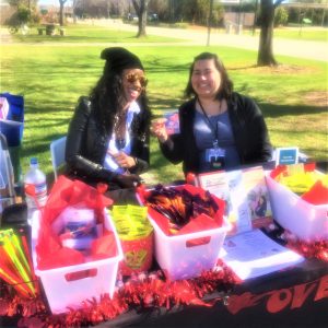 Elicia Stewart, Harmony Health Community Events Coordinator smiles and laughs while Christina Villegas, Outreach Enrollment Specialist holds up a strawberry dental dam.
